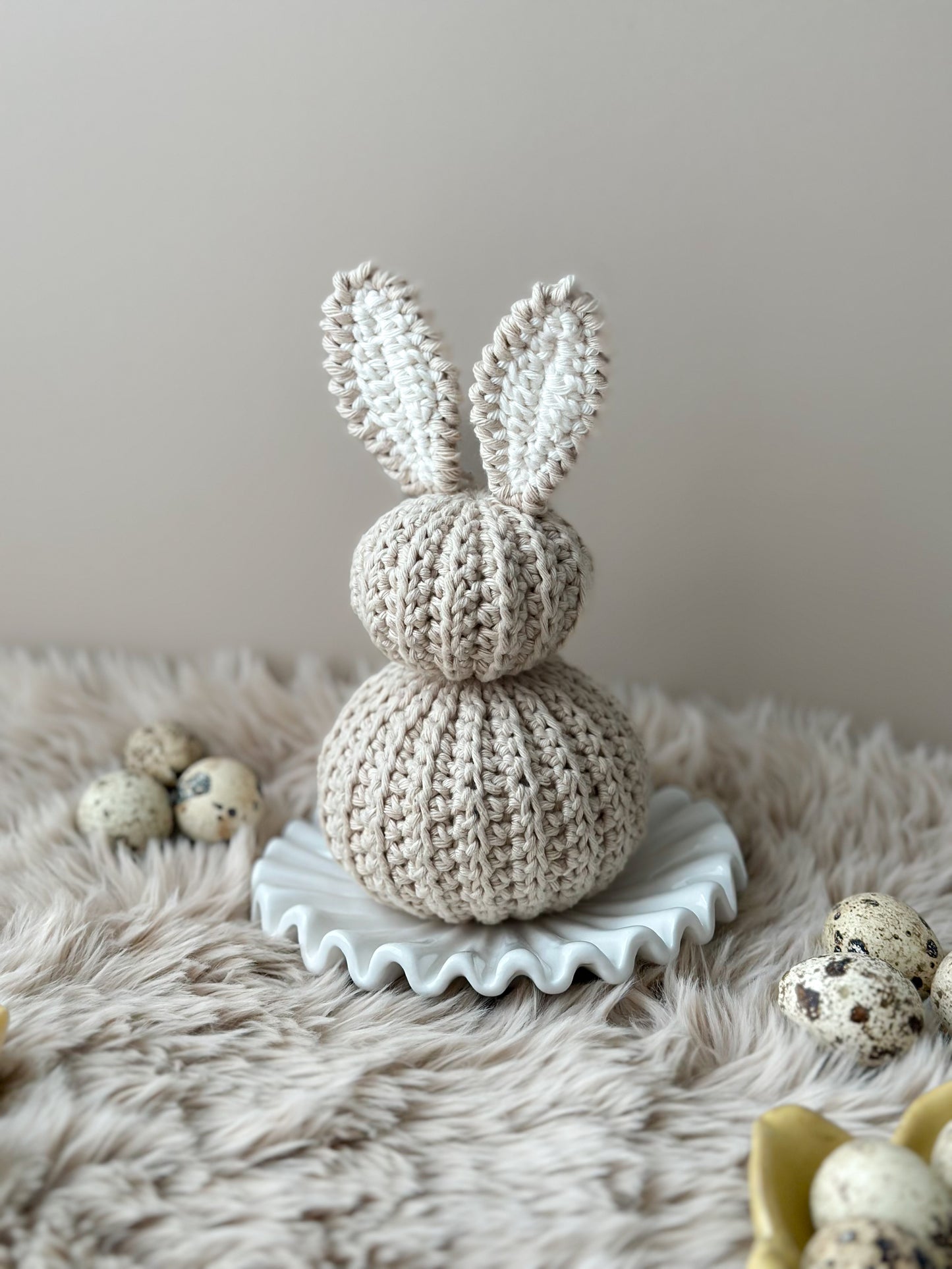 Crocheted  bunny figure on a decorative stand with small eggs on a fur surface. A Crocheted nursery room decor or crocheted easter decoration. Crochet pastel bunnies. Babyshower gift. Handmade gift. Animo Design. Animodesignshop.