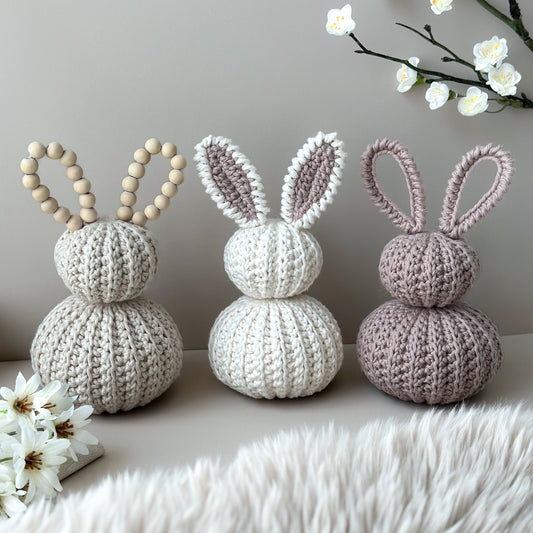 Three crocheted bunnies in different colors on a white surface with flowers in the background. Crocheted nursery room decor or crocheted easter decoration. 