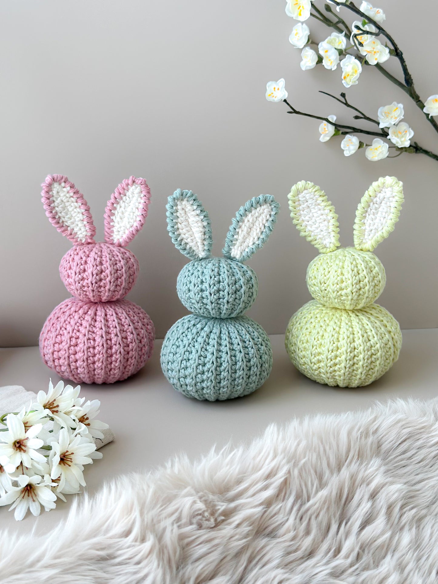 Three crocheted bunny figures on a light surface with flowers in the background. Crocheted nursery room decor or crocheted easter decoration. Crochet pastel bunnies. Babyshower gift. Handmade gift. Animo Design. Animodesignshop.