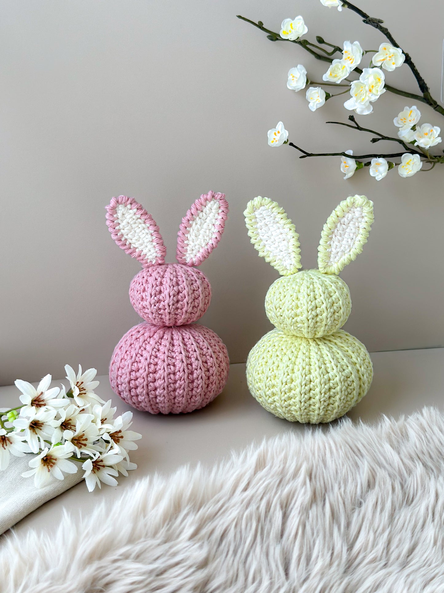 Two crocheted bunny figures on a light surface with flowers in the background. Crocheted nursery room decor or crocheted easter decoration. Crochet pastel bunnies. Babyshower gift. Handmade gift. Animo Design. Animodesignshop.