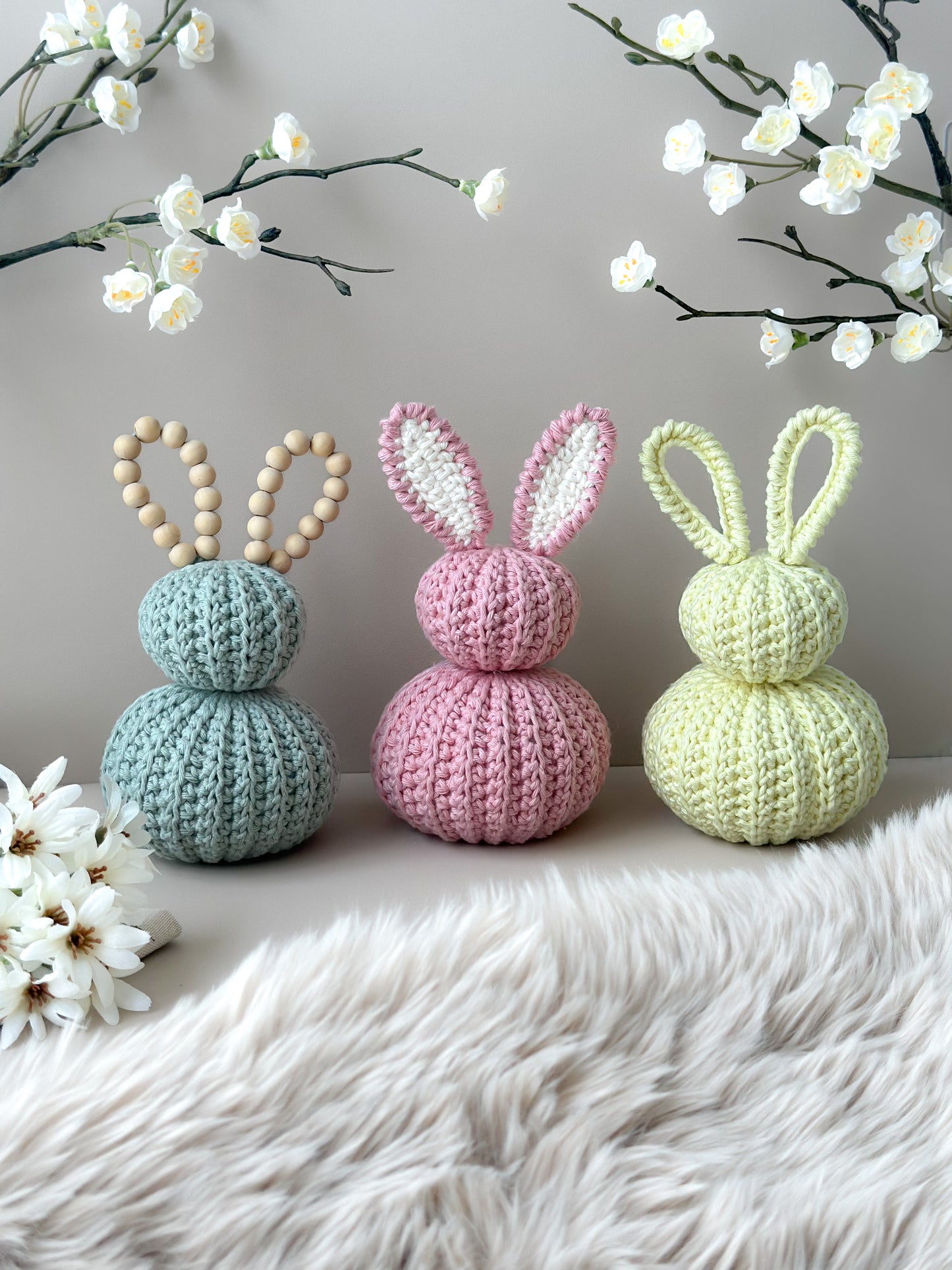 Three crocheted bunny figurines in pastel colors on a soft surface with floral decorations. Crocheted nursery room decor or crocheted easter decoration. Crochet pastel bunnies. Babyshower gift. Handmade gift. Animo Design. Animodesignshop.