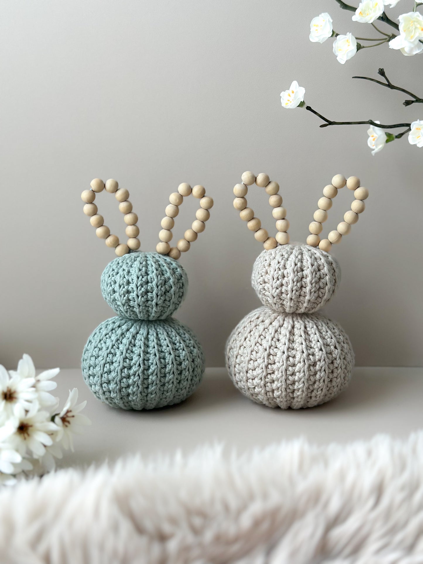 Two crocheted bunny figures with wooden ears on a light surface with flowers in the background. Crocheted nursery room decor or crocheted easter decoration. 