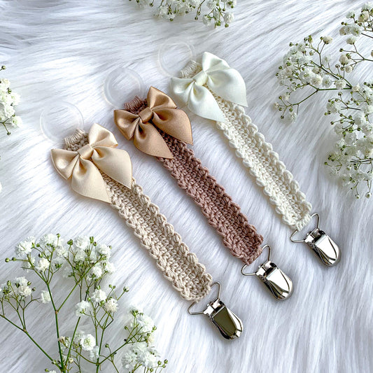 Three crocheted baby pacifier clips with satin bows in beige, blush pink, and cream, arranged on a white faux fur background with white baby's breath flowers.