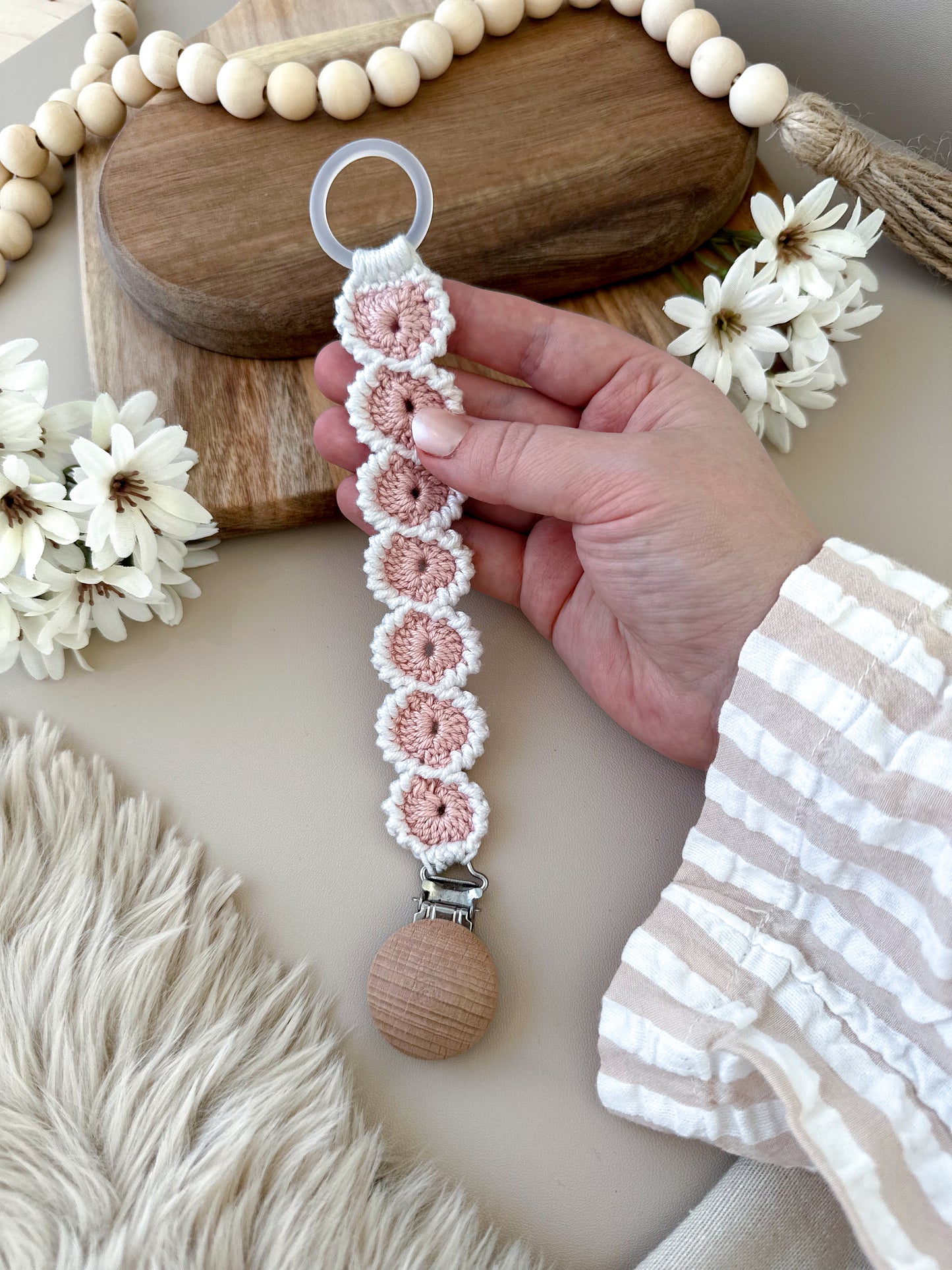 En hand som håller upp ett rosa virkat nappband, som visar det detaljerade hantverket. Svensk design för de allra minsta