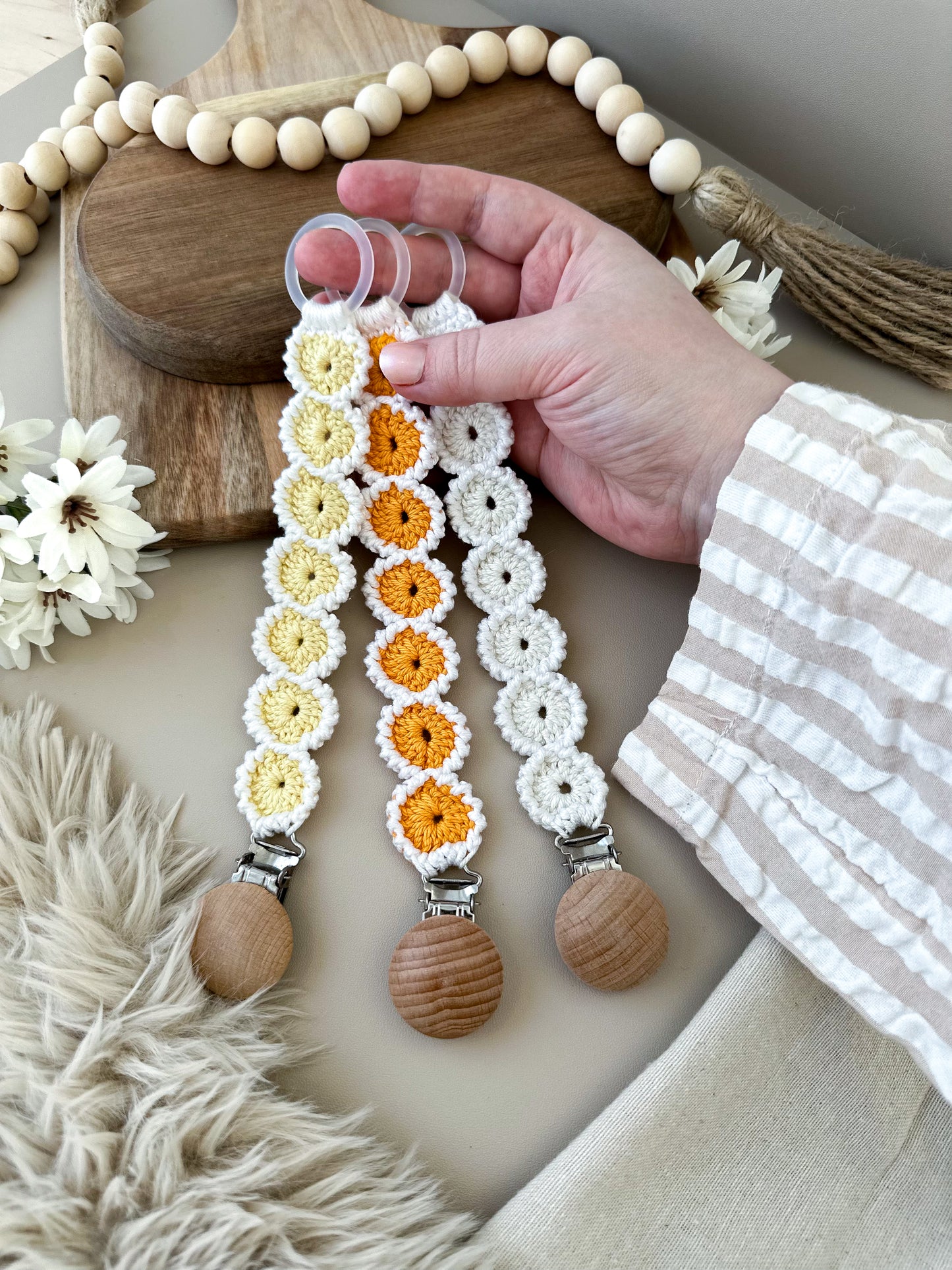Hand håller tre virkade napphållare i ljus, skandinavisk blommig design. Handgjorda babytillbehör i naturliga och varma färgtoner.